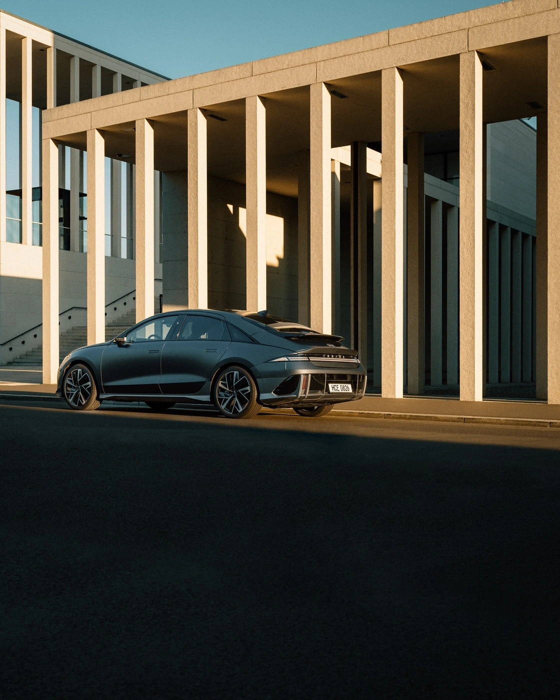 A sleek grey electric sedan parked outside a monumental building with striking concrete columns under warm daylight.