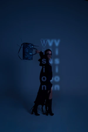 A woman in a black outfit wearing sunglasses holds up a dark handbag against a blue studio background with projected text.