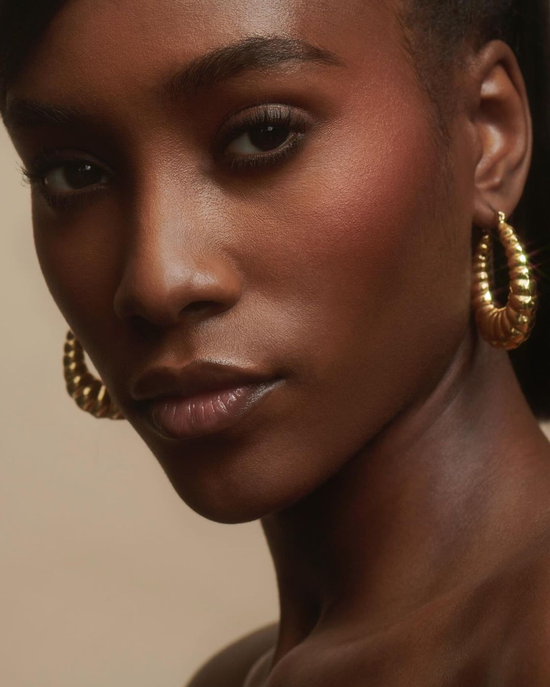 Close-up portrait of an African American woman with a serious expression, wearing gold hoop earrings, against a beige background.