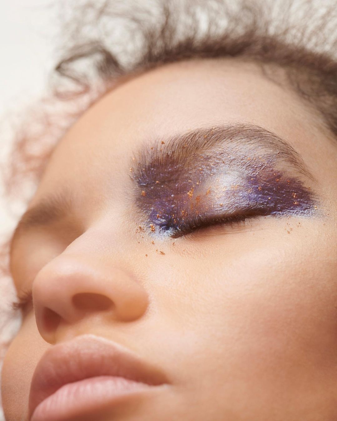 Close-up of a woman's face with striking makeup highlighting her eyebrow and eyelid in shades of purple and gold.