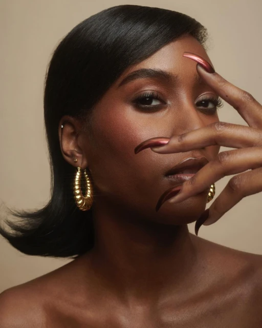 Close-up portrait of an African American woman with dark hair and shiny dark-red nails, wearing gold hoop earrings against a tan background.