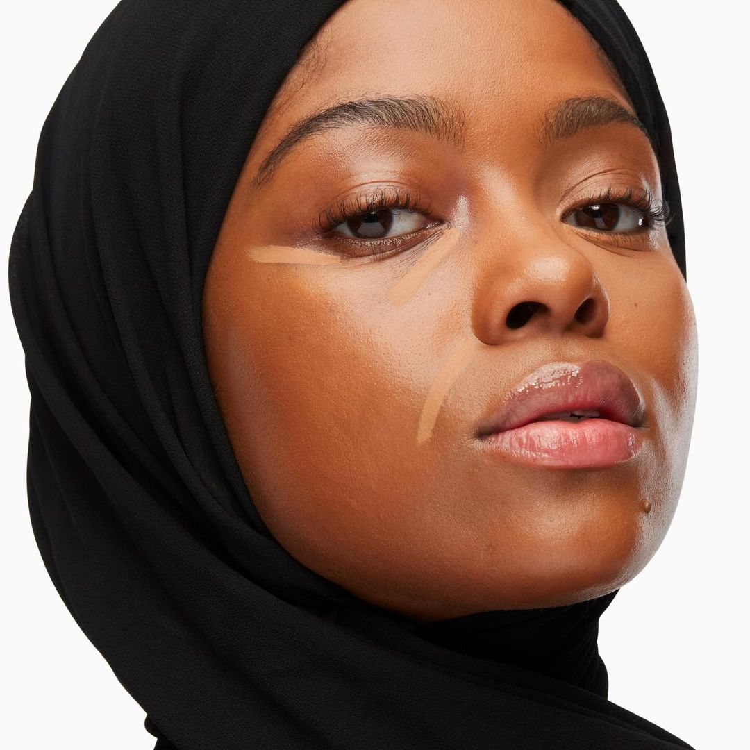 Close-up of a woman with tan skin wearing a black headscarf, and makeup, set against a clean white backdrop in a studio setting.