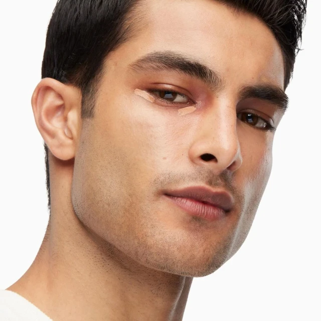 Close-up of a man applying concealer under his eye; beauty and personal care concept for cosmetics and skincare product advertising.