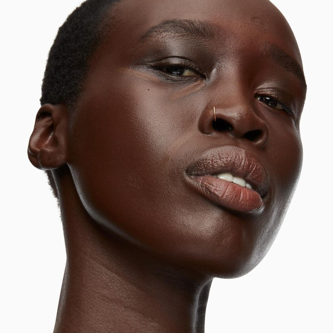 Close-up portrait of a dark-skinned adult woman with a nose ring, natural makeup, and shiny skin, against a plain white background.