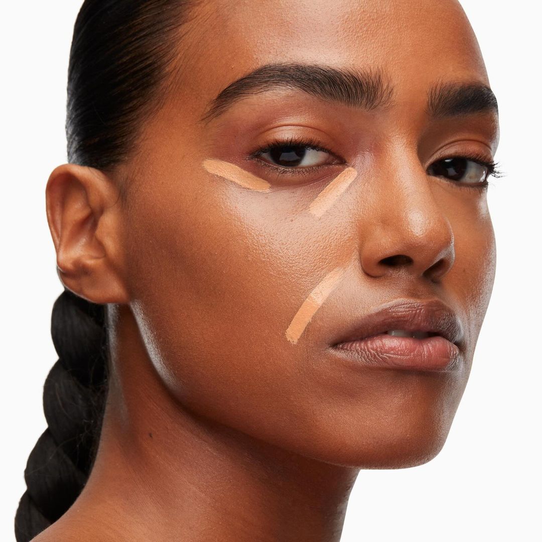 Close-up of a dark-skinned woman's face with tan concealer applied under her eyes and on her cheek, against a white background.