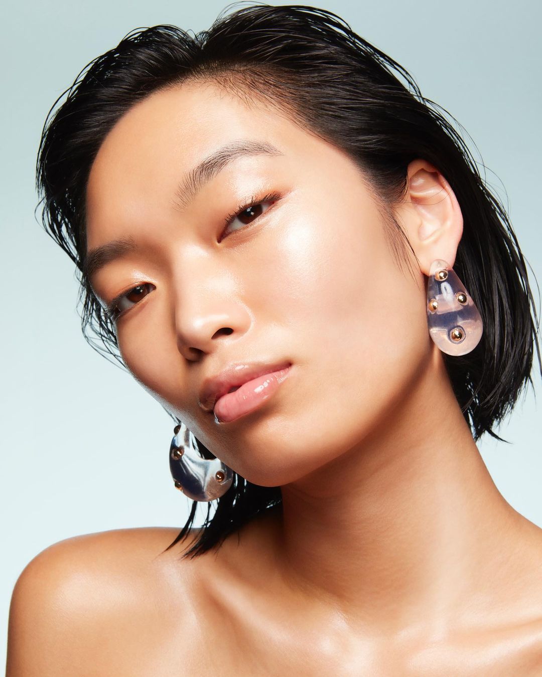 Close-up studio shot of a young adult woman with wet dark hair, shiny skin, and large earrings on a pale blue backdrop.