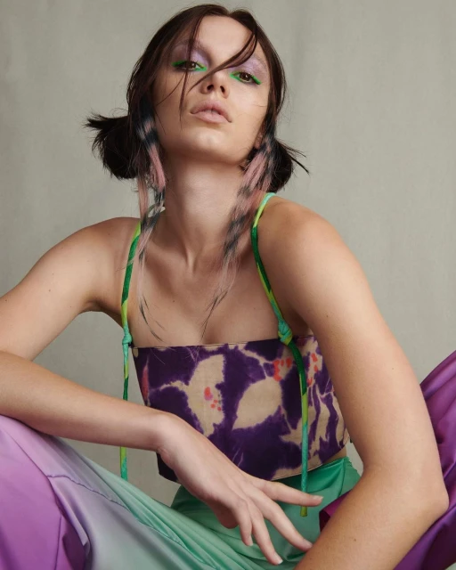 A young woman with decorated pigtails and dramatic makeup poses indoors wearing a floral top and gradient pants in a fashion portrait.