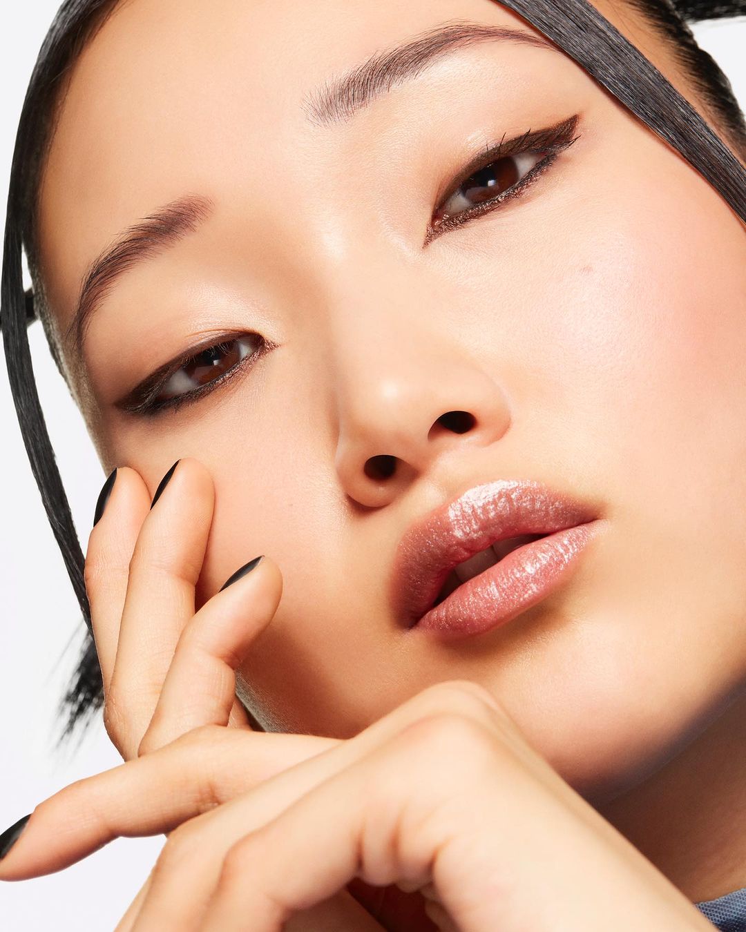 Close-up of a young Asian woman with dramatic eyeliner and glossy lips, posing with her hand near her face, set against a white background.
