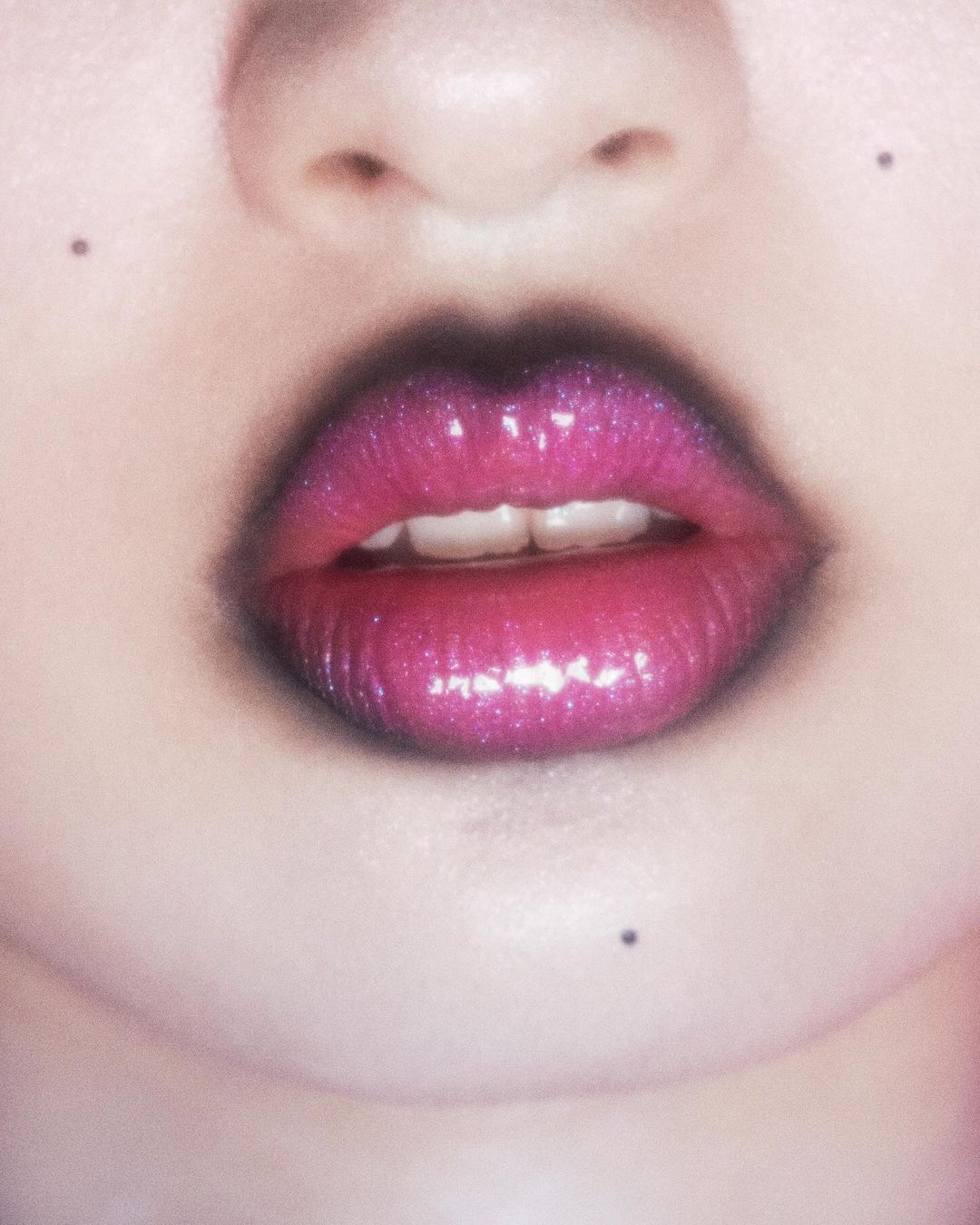 Close-up of a woman's face featuring pink and purple glitter lipstick, fair skin, and visible details like pores and a beauty mark.