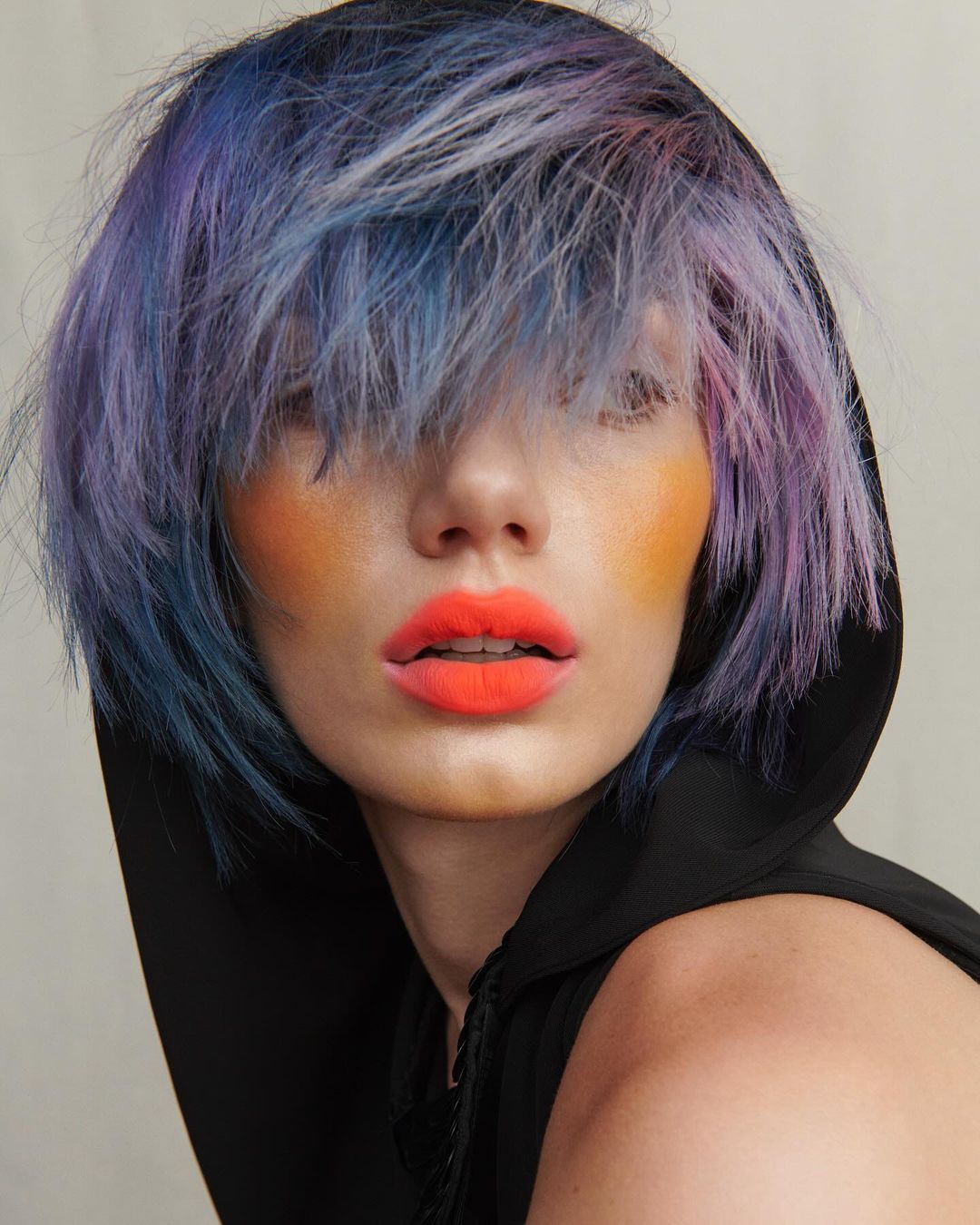 Close-up portrait of a woman with vivid blue and purple hair, bright orange lipstick, and orange makeup on her cheeks wearing a black hood.