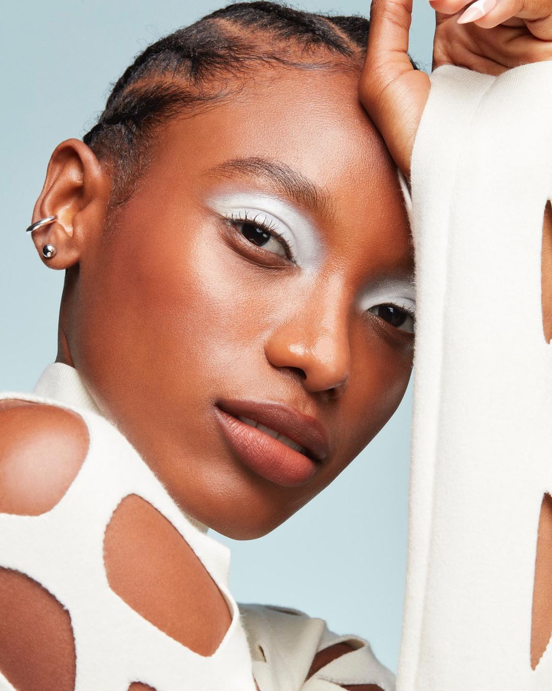 Close-up studio shot of a young African American woman with white eyeshadow and a white cut-out sweater on a light blue background.