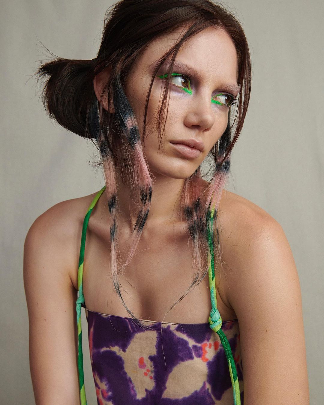 Portrait of a young woman with pink-streaked hair, wearing colorful eye makeup, and a patterned top against a neutral backdrop.