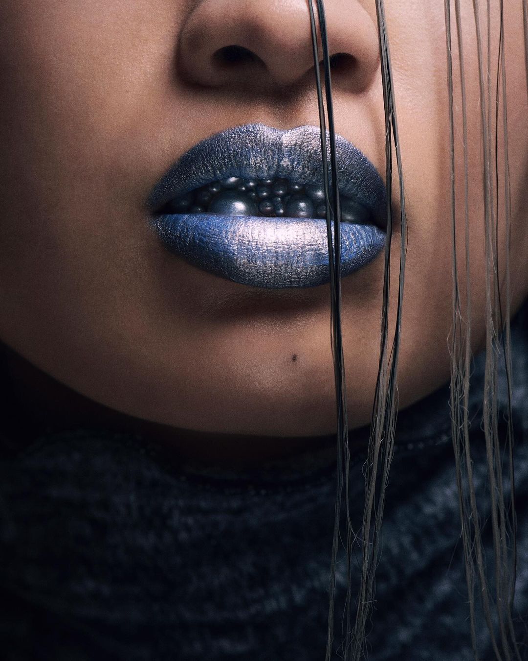 Close-up portrait of an adult woman with a dark complexion, wearing dark blue lipstick and black hair strands over her face.