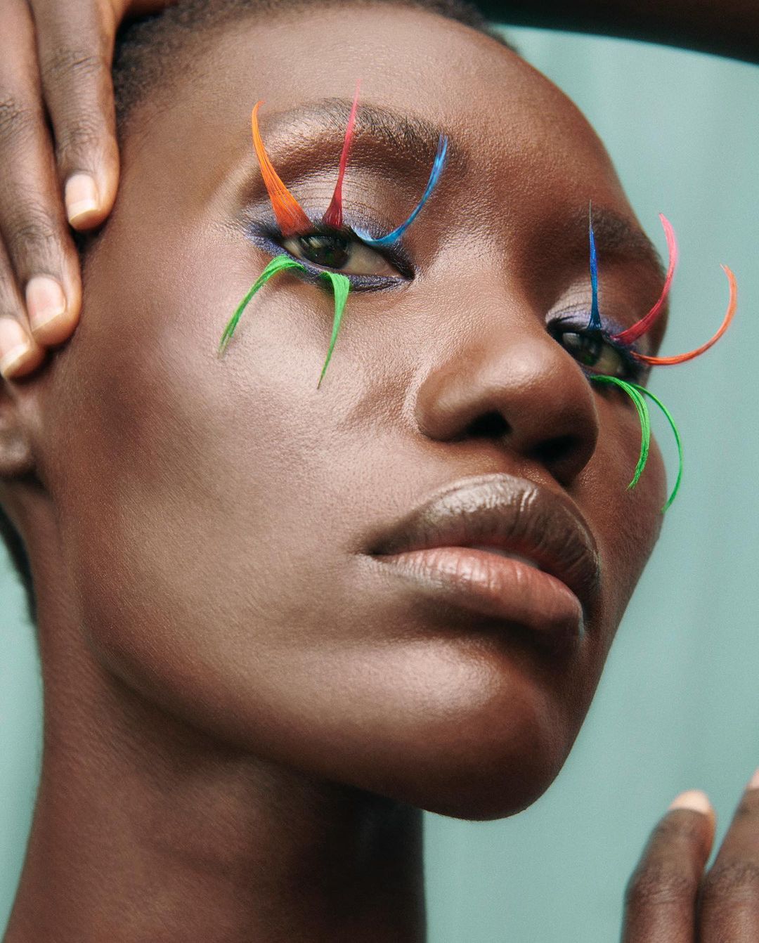 Close-up portrait of a woman with creative rainbow eye makeup and dark skin in a studio with hands framing her face.