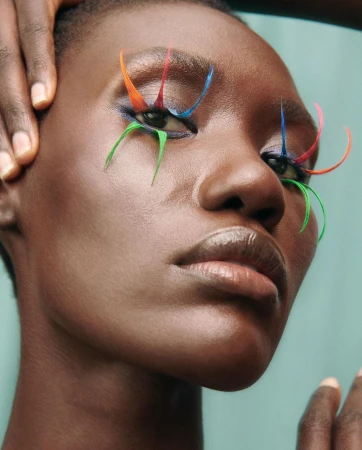 Close-up portrait of a woman with creative rainbow eye makeup and dark skin in a studio with hands framing her face.