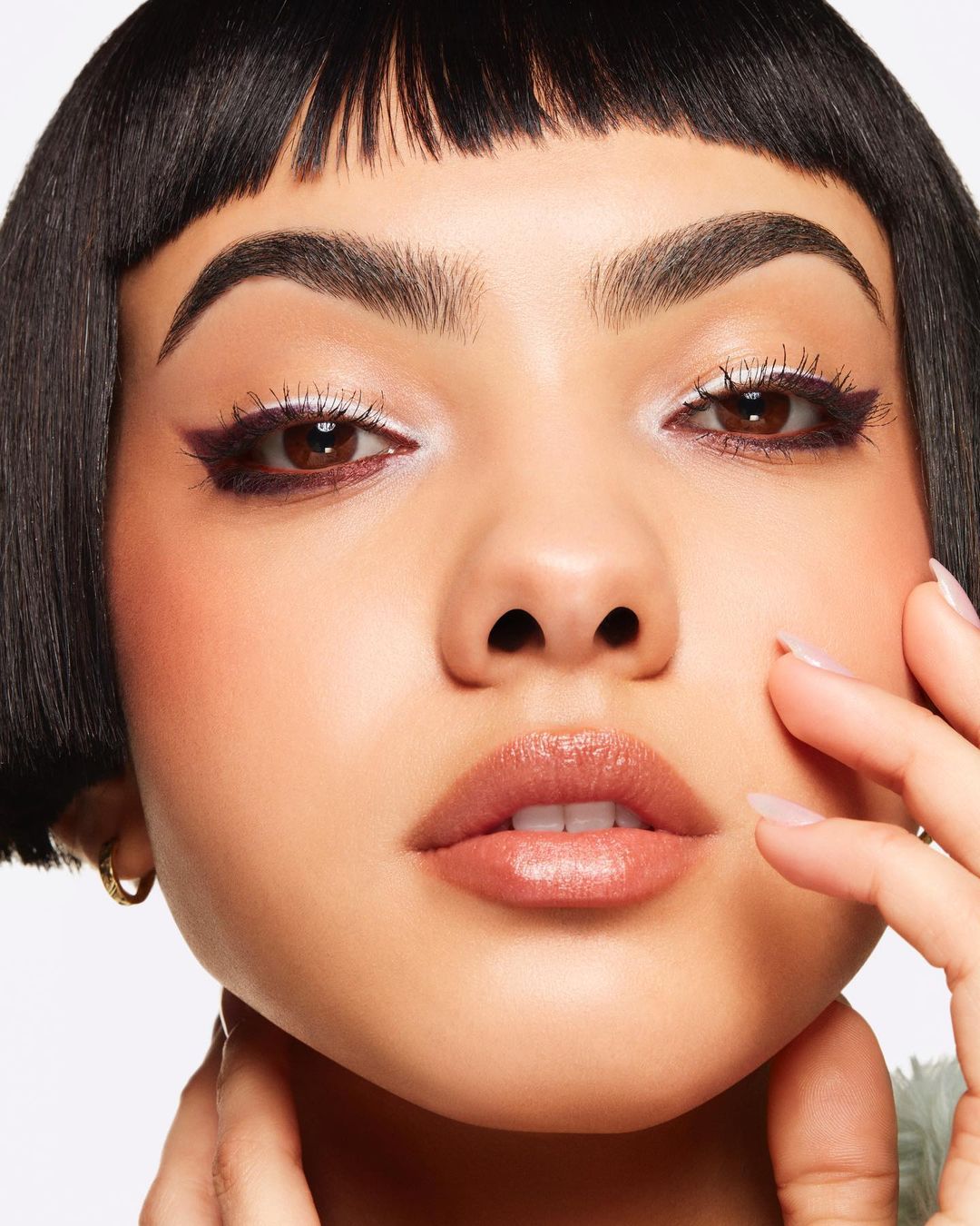 Close-up portrait of a woman with dark hair and light skin, wearing unique eye makeup, against a plain white backdrop in a beauty context.