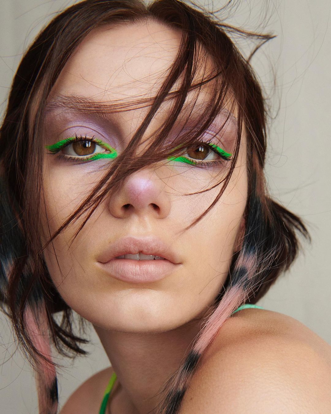 Close-up of a young adult woman with dark brown hair and brown eyes wearing lilac eyeshadow and bright green eyeliner in a studio setting.