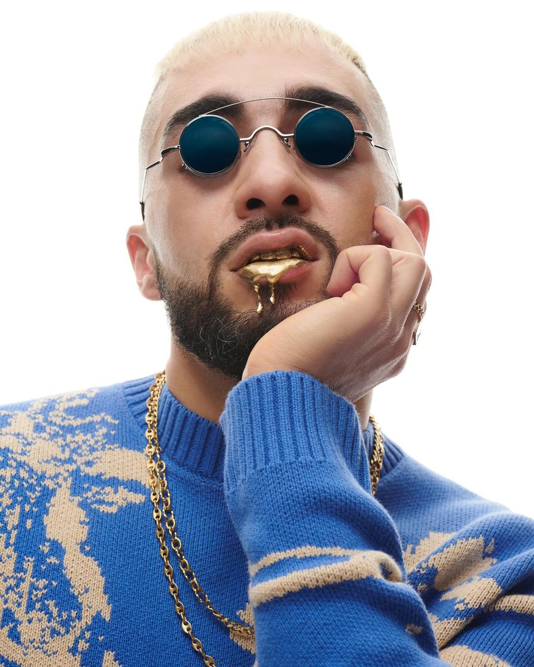 Portrait of a male musician wearing round sunglasses, a patterned blue sweater, a thick gold chain, and gold teeth set against a white background.