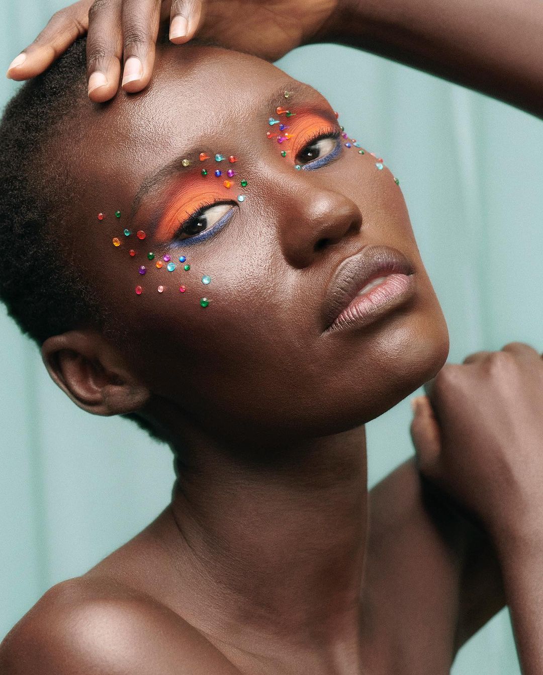 Close-up portrait of a dark-skinned female model with colorful rhinestones on her face and orange eyeshadow against a light teal background.