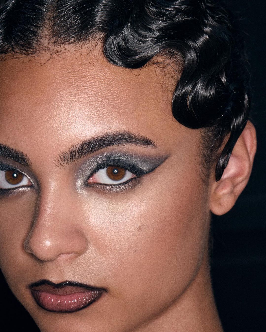 Close-up portrait of a woman with dramatic eye makeup and glossy lips in a dimly lit setting, showcasing beauty and cosmetic art.