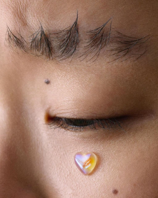 Close-up of a person's face with striking eyebrows and a heart-shaped rhinestone on their cheek, capturing unique beauty details.