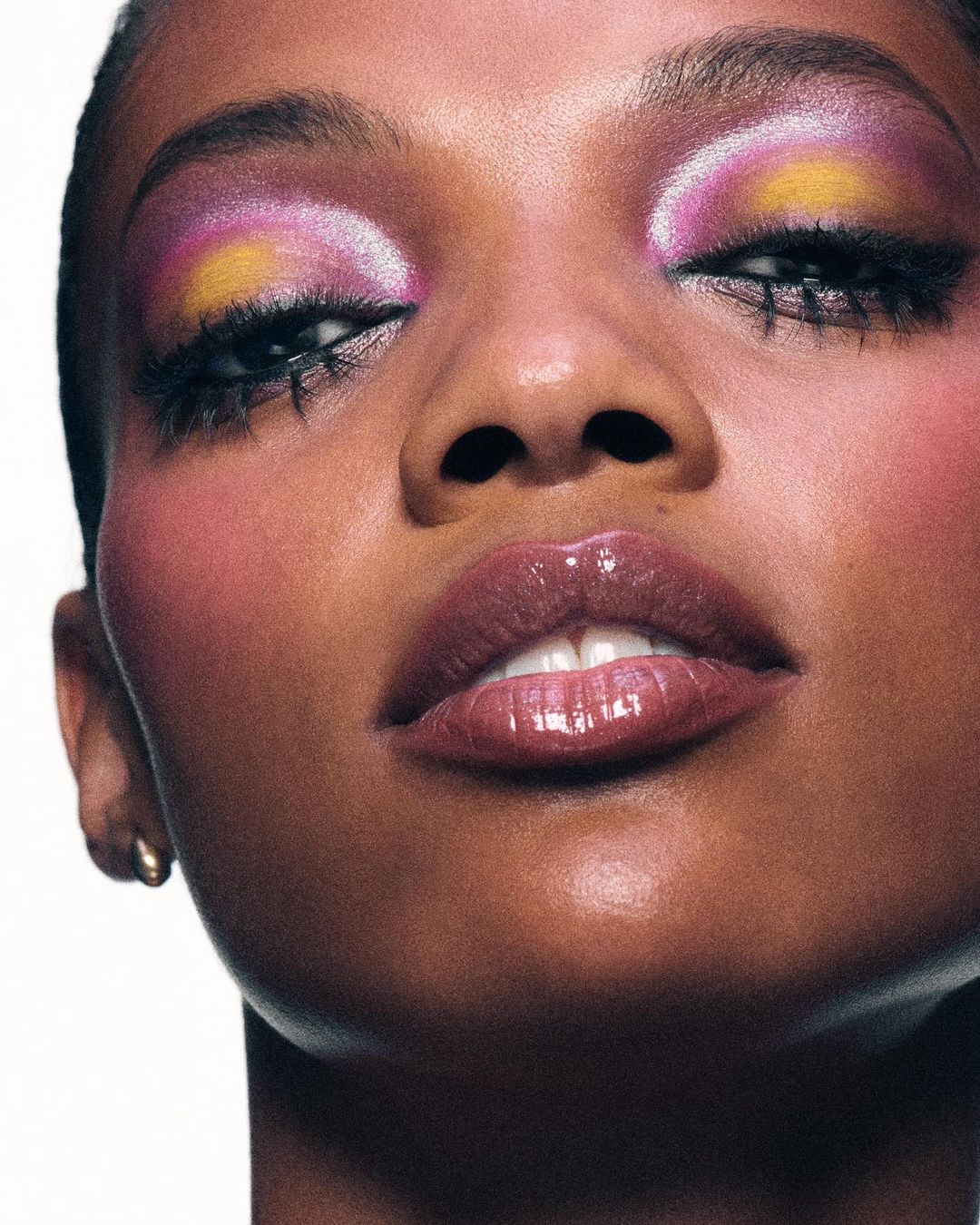 Close-up shot of a dark-skinned woman showcasing dramatic yellow, pink, and purple eye makeup, paired with glossy lips against a clean, white backdrop.