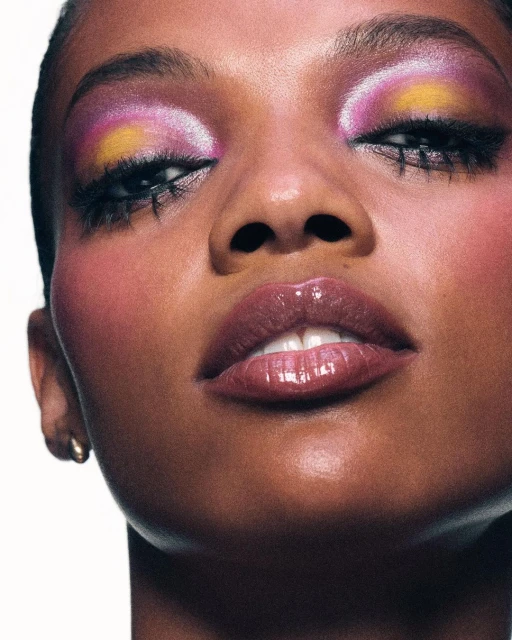 Close-up shot of a dark-skinned woman showcasing dramatic yellow, pink, and purple eye makeup, paired with glossy lips against a clean, white backdrop.