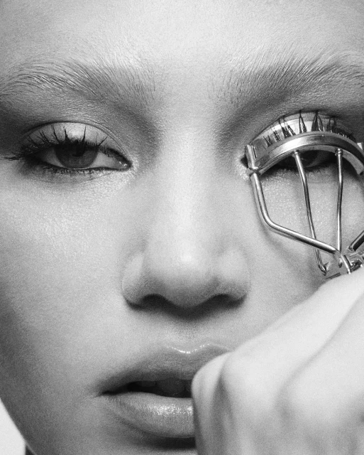 Close-up black and white shot of a fair-skinned adult female using a metal eyelash curler, focusing on beauty and eye makeup.