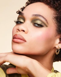 Close-up portrait of a woman with green eyeshadow, nude lips, and gold hoop earrings resting her chin on her hands.