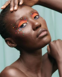 Close-up portrait of a Black woman with colorful rhinestones around her eyes and vibrant orange eyeshadow, photographed by Marion Clémence Grand