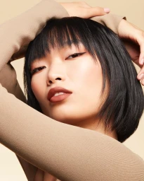Close-up portrait of a woman with dark bob hair and winged eyeliner posing with her hands behind her head against a beige background.