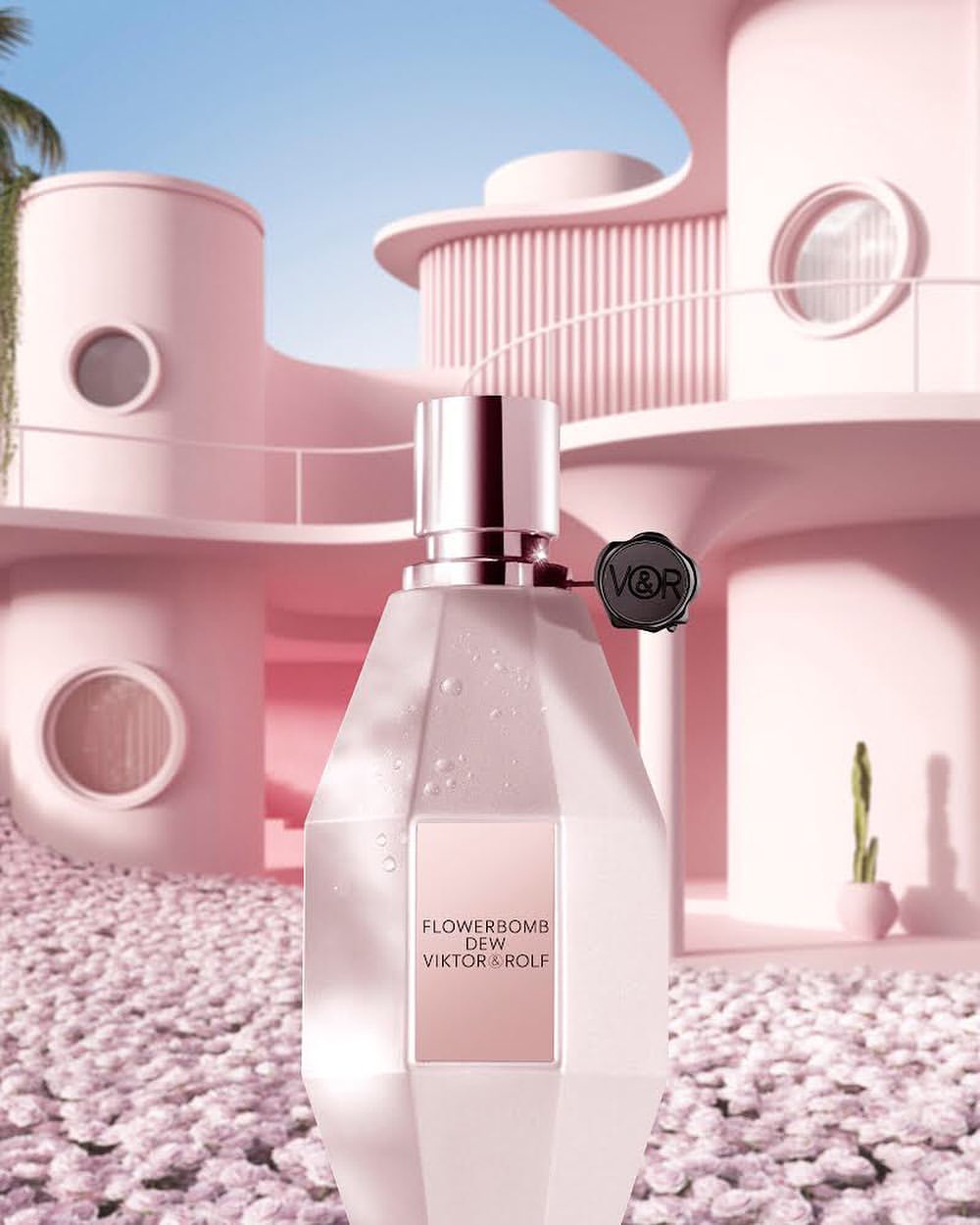 Crystal-shaped Viktor&Rolf Flowerbomb Dew perfume bottle stands out against a soft pink building and white pebble ground.