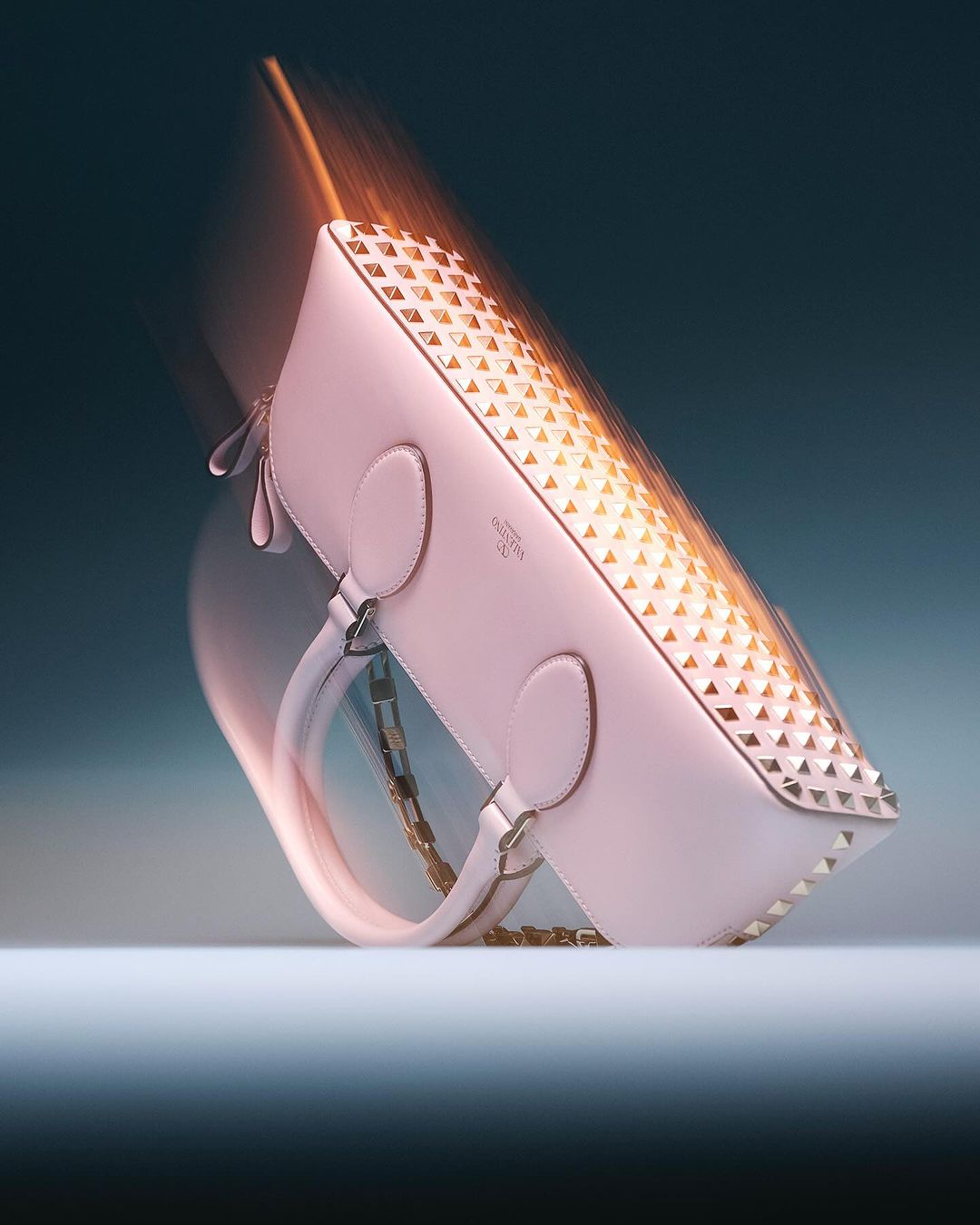 Elegant pink leather handbag with silver stud details, gold chain, and motion blur on a gradient blue background in a studio shot.