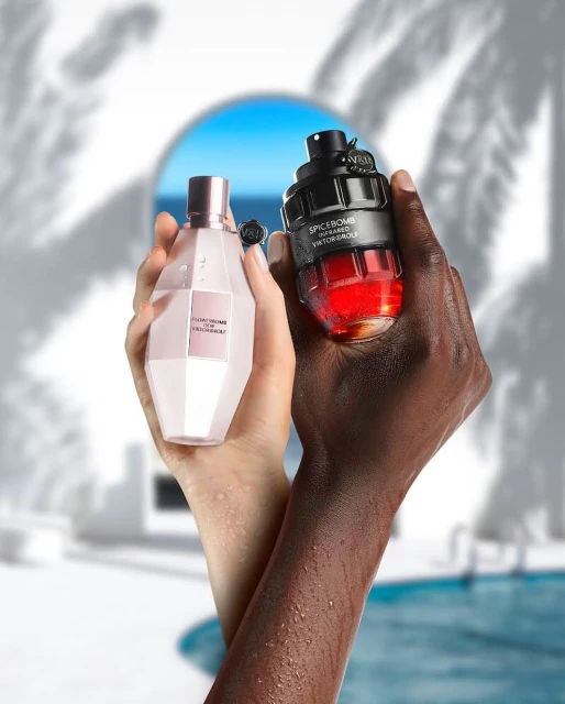 Promotional shot of two perfume bottles, Flowerbomb and Spicebomb, held in the hands of adult male and adult female with beach background.