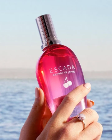 Hand holding a bright pink Escada perfume bottle with cherry graphic against a sparkling ocean background.