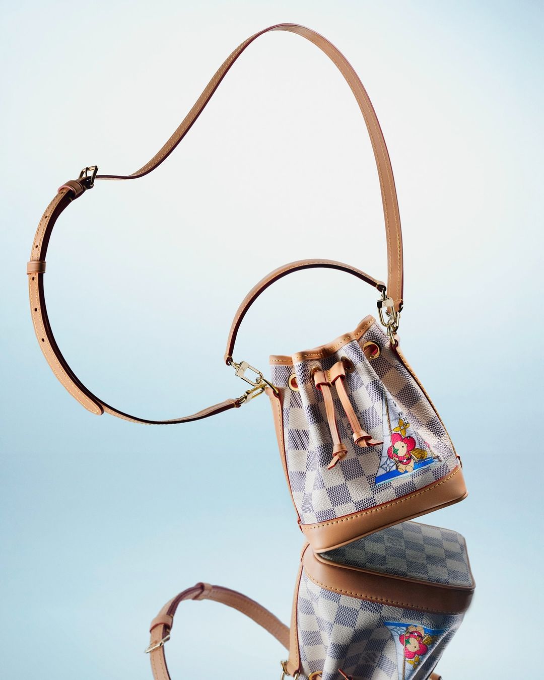 Upscale bucket bag featuring a checkerboard pattern and whimsical cartoon character graphic, paired with a tan leather strap on a light gradient.