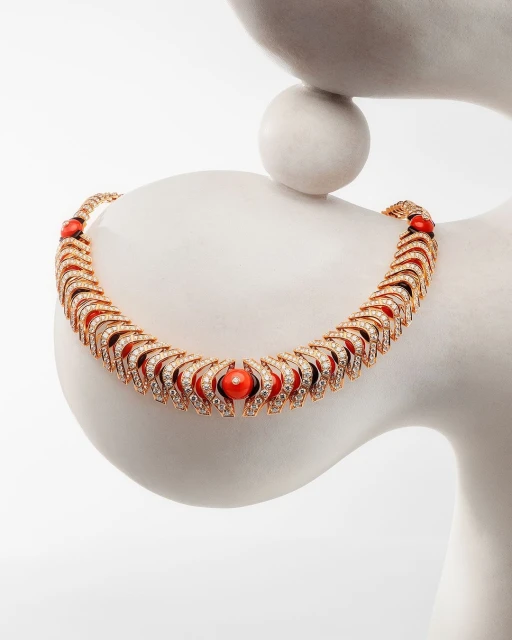 Elegant gold and diamond necklace with coral beads displayed on a white abstract sculpture against a clean, bright white backdrop.