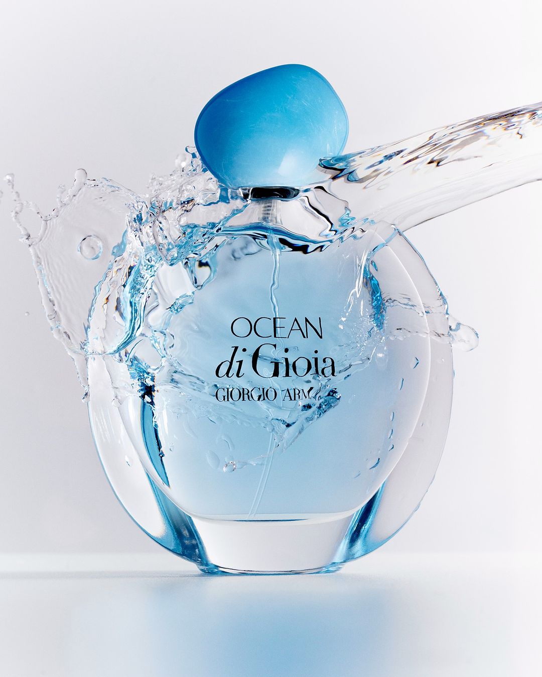 Close-up of a clear, round Giorgio Armani Ocean di Gioia perfume bottle with a blue cap, surrounded by a splash of clear water against a white background.