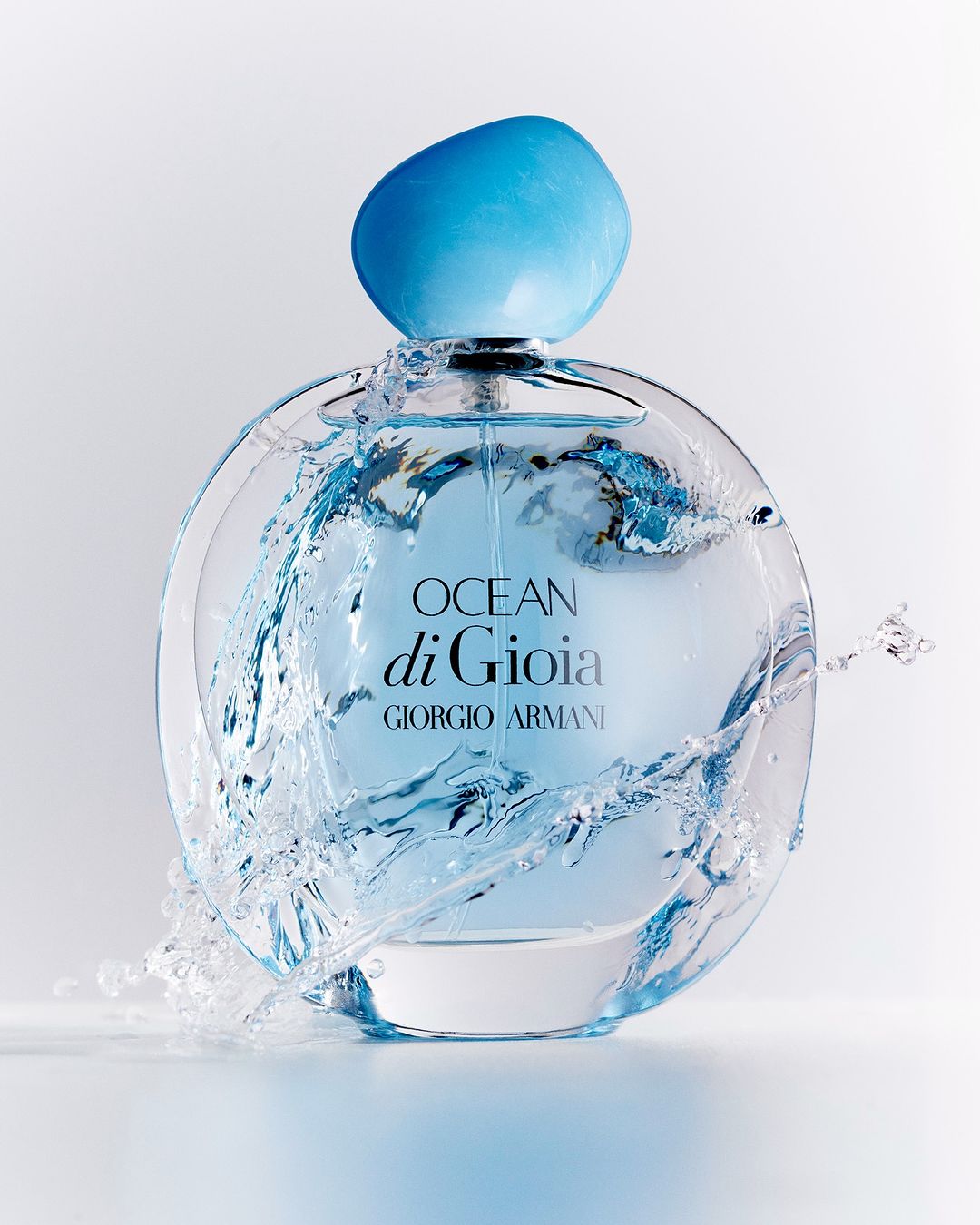 Giorgio Armani Ocean di Gioia perfume bottle, clear glass with a light blue cap, surrounded by splashing water against a white background.