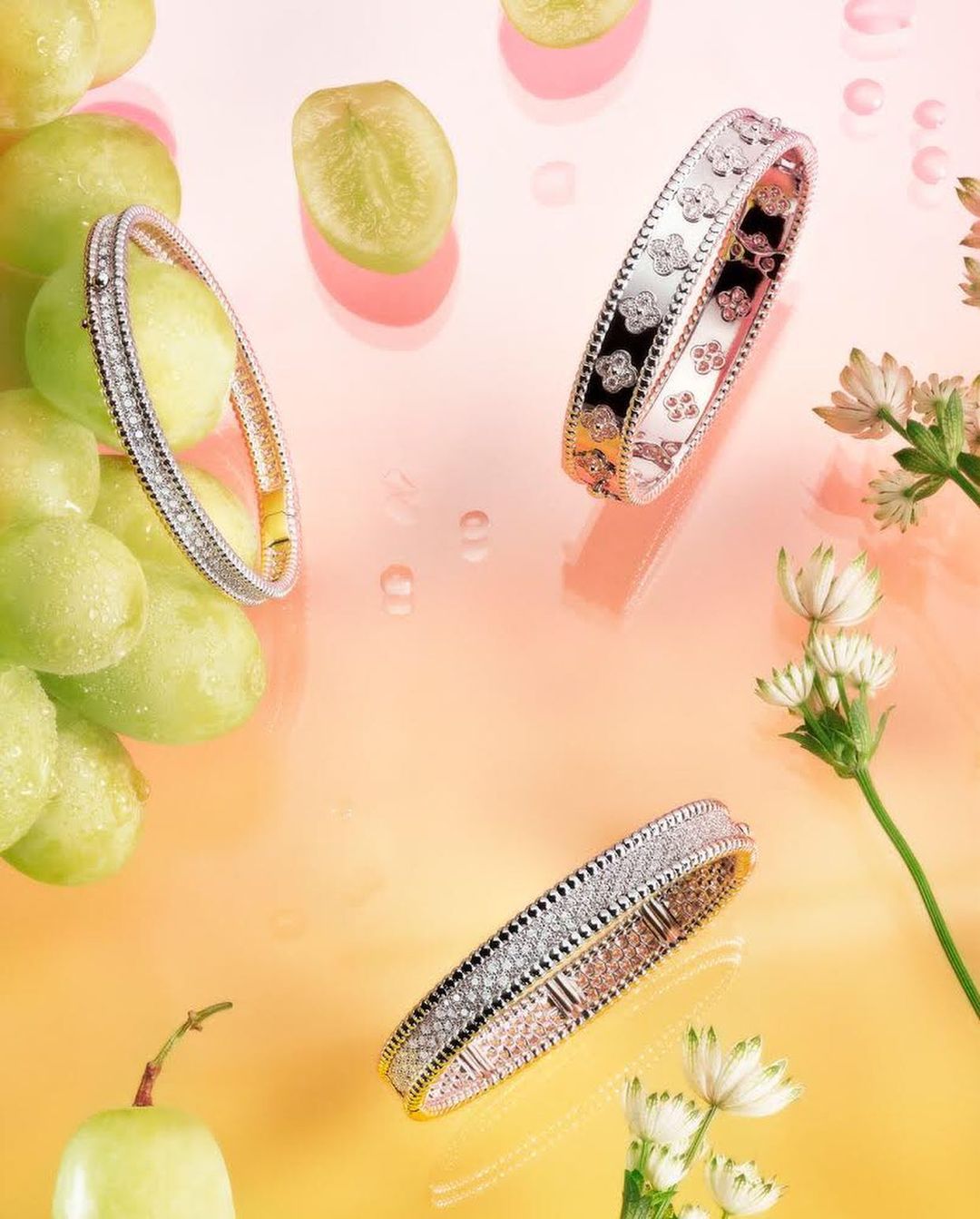 Three diamond bangles are showcased with green grapes and white flowers against a gradient peach and yellow background, high angle view.