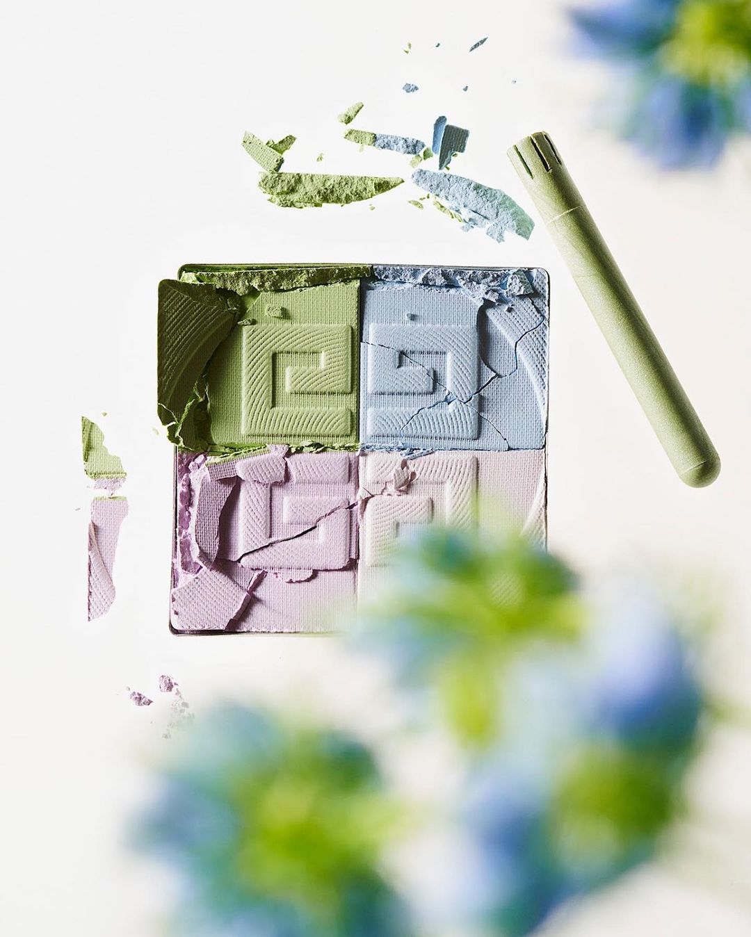 Overhead view of an open, four-color eyeshadow palette with a double-ended applicator and scattered powder remnants on a white surface.