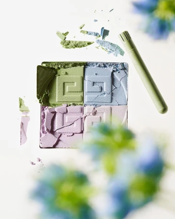 Overhead view of an open, four-color eyeshadow palette with a double-ended applicator and scattered powder remnants on a white surface.