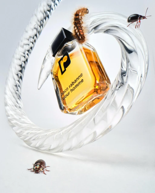 Close-up product shot of a Paco Rabanne bottle with a caterpillar and beetle on it, set against a white background.