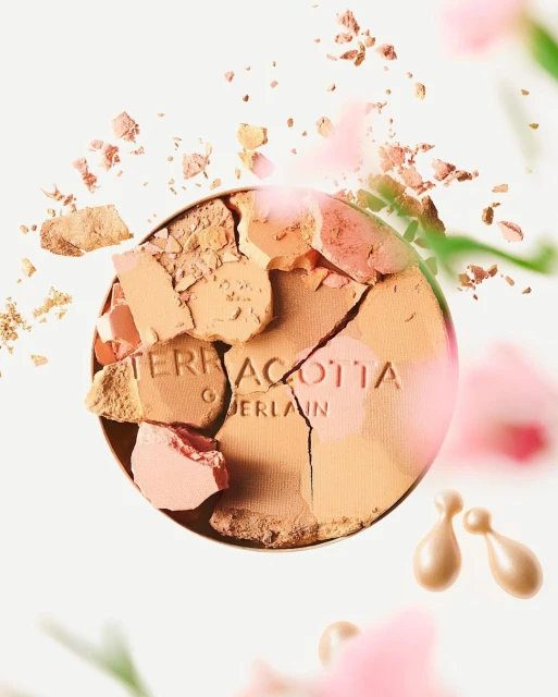Close-up studio shot of cracked Guerlain Terracotta bronzing powder with scattered fragments and a few pearl skincare capsules around it.