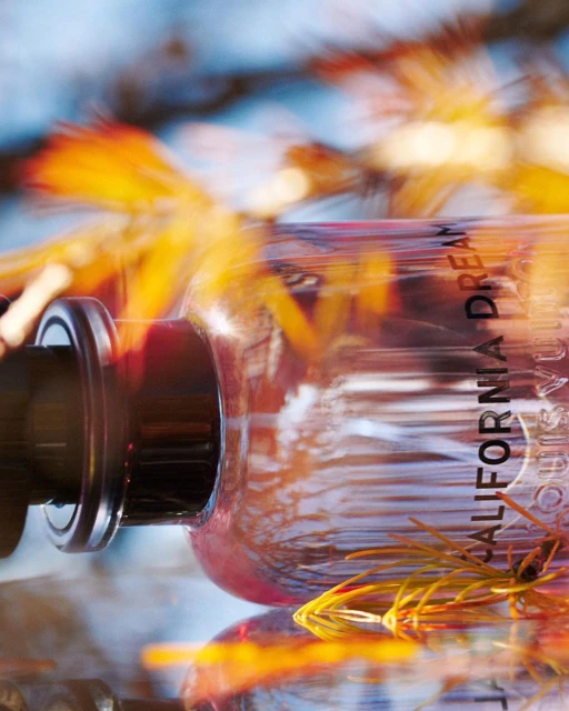 Perfume bottle on a reflective surface with orange and yellow floral accents in a blurred background, product photography.