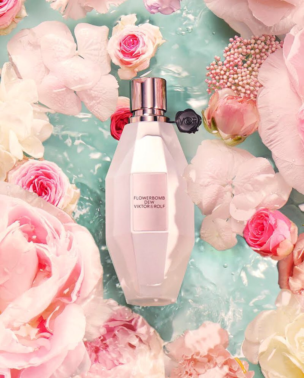 Flowerbomb Dew Viktor&Rolf fragrance bottle surrounded by pink and white flowers floating in light blue water, creating a serene beauty scene.