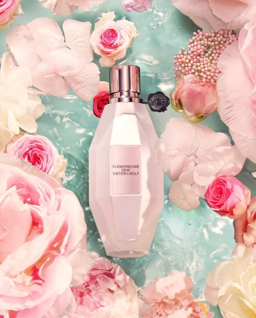 Flowerbomb Dew Viktor&Rolf fragrance bottle surrounded by pink and white flowers floating in light blue water, creating a serene beauty scene.