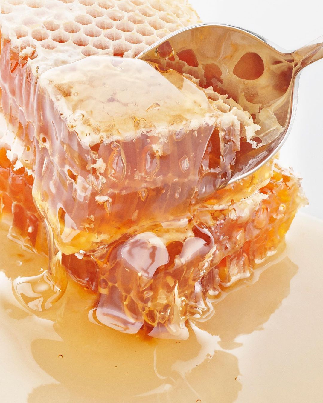 Close-up of honeycomb pieces on a white surface, drizzled with honey, next to a silver spoon. Food and beverage photography.