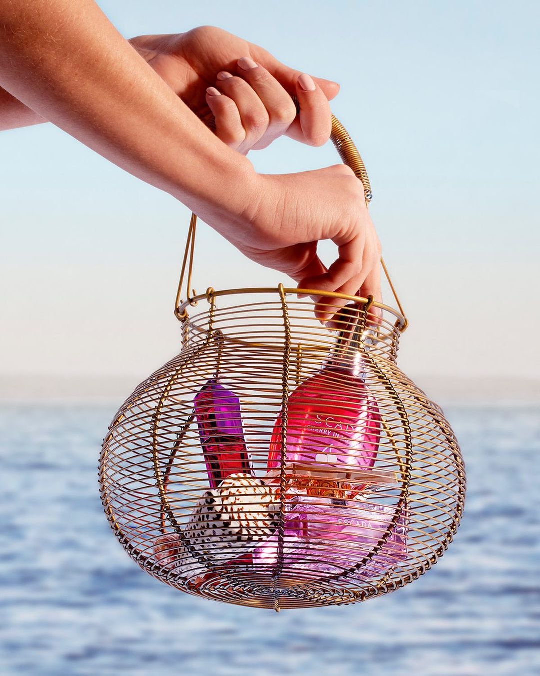 Close-up shot of a person's hands holding a gold wire basket containing two glass bottles against a blue sky and water backdrop.