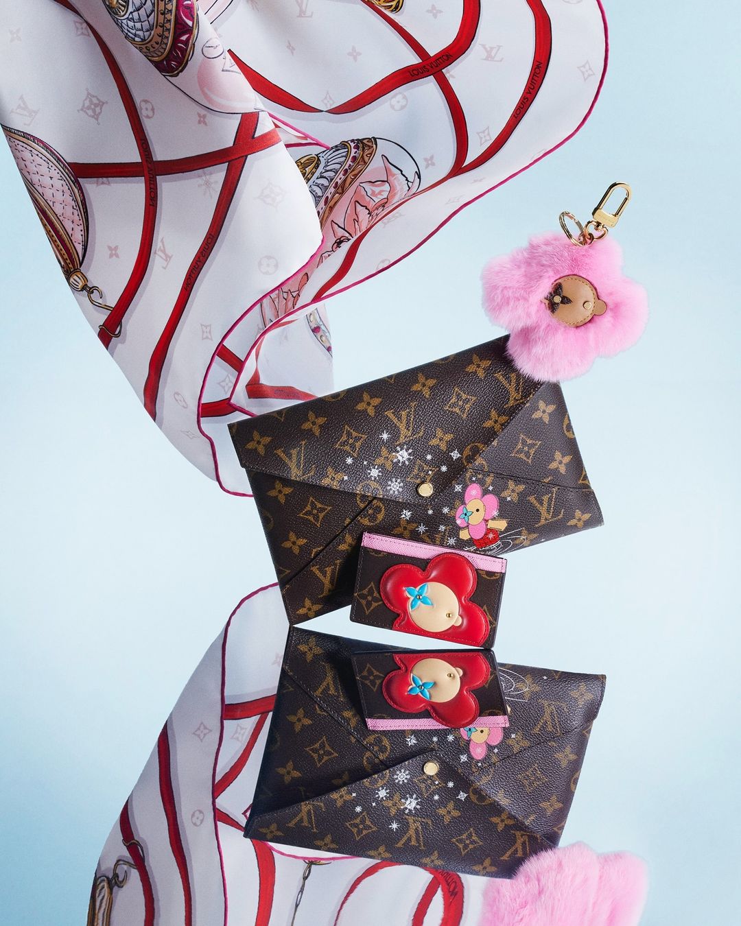 Fashion layout featuring floating monogrammed envelope bags, a pink furry keychain, and a printed silk scarf against a light blue background.